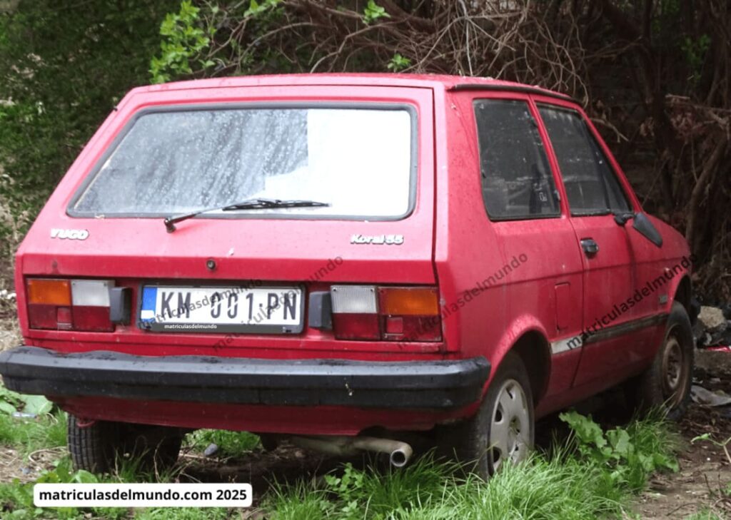 Yugo Koral rojo con matrícula KM de Kosovo estacionado bajo la lluvia.